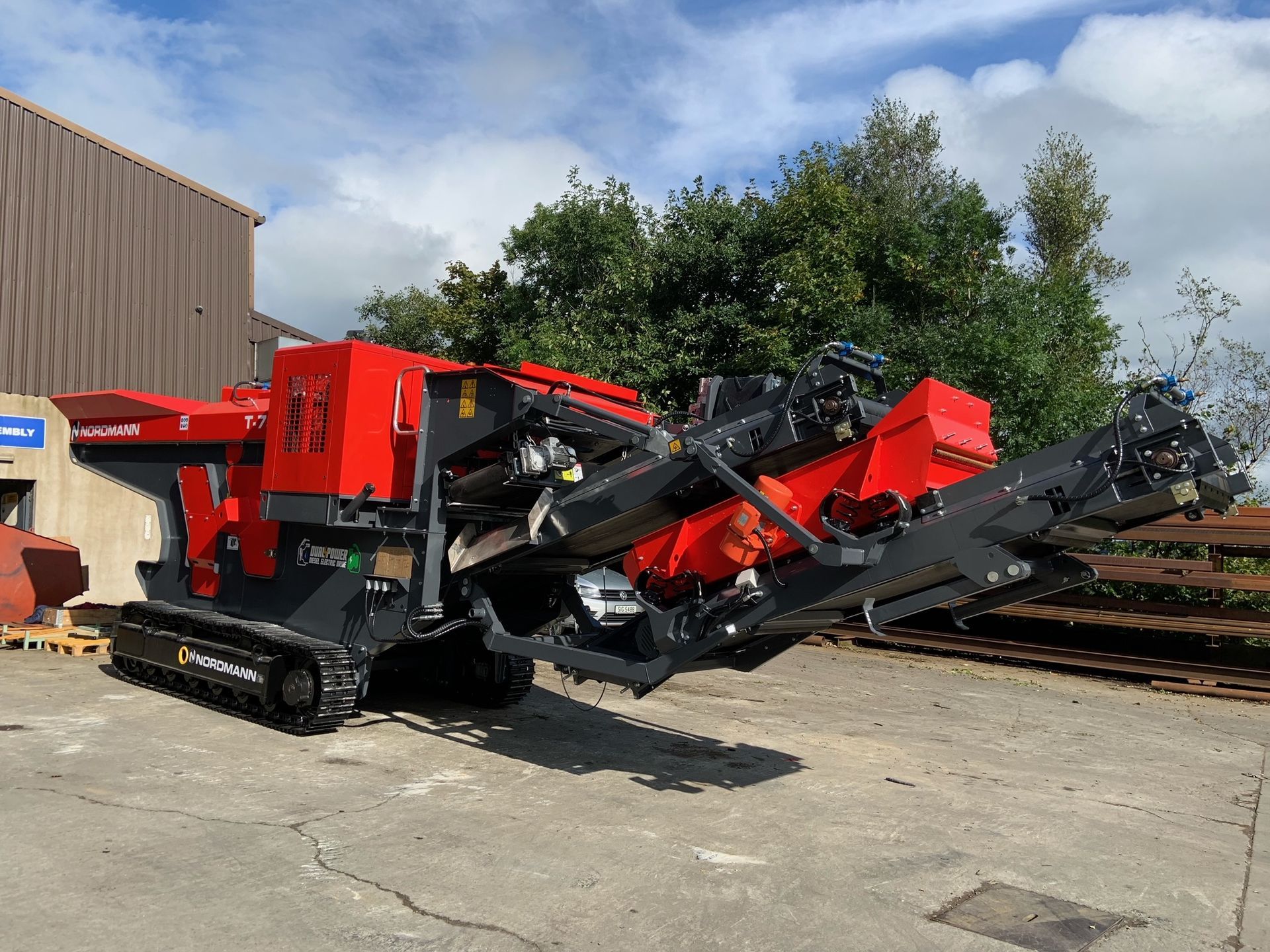 Red and black mobile rock crusher on tracks outdoors.  A machine with extended conveyor belts in front of a building and trees.