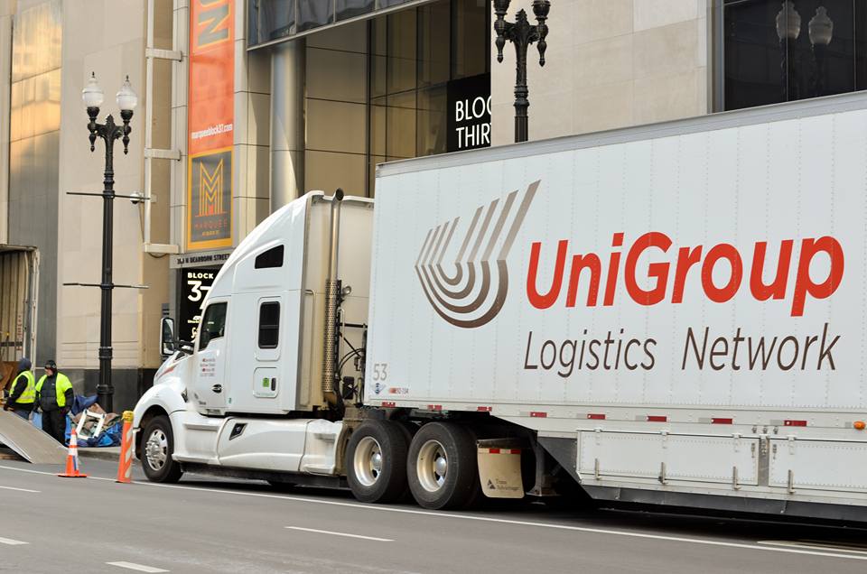 UniGroup Logistics truck parked by a city building, with workers unloading nearby