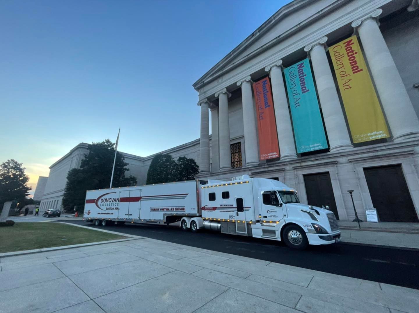 Moving truck parked beside a neoclassical building with tall columns and colorful banners
