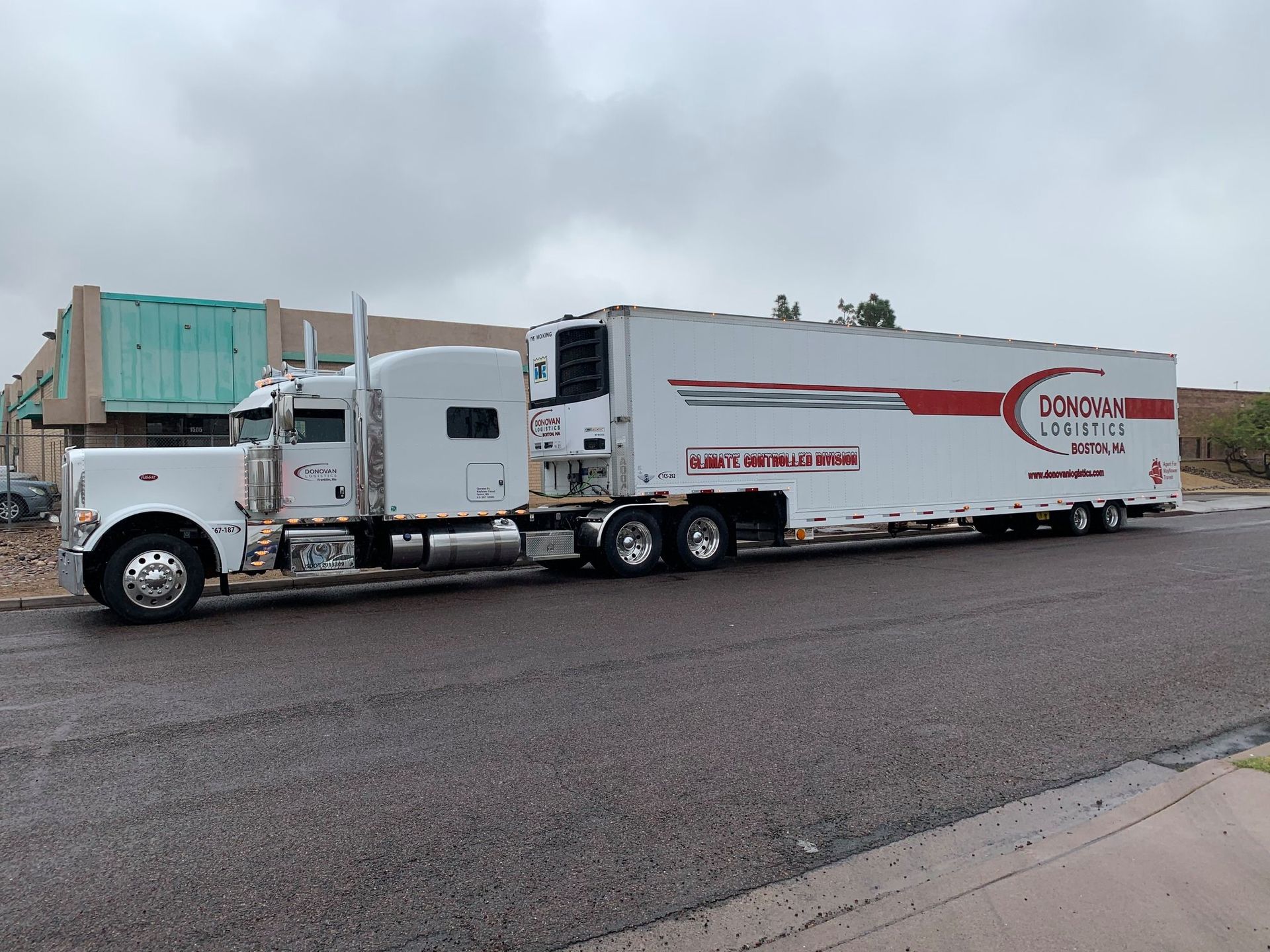 White semi-truck with a long trailer parked on a wet lot under cloudy skies.