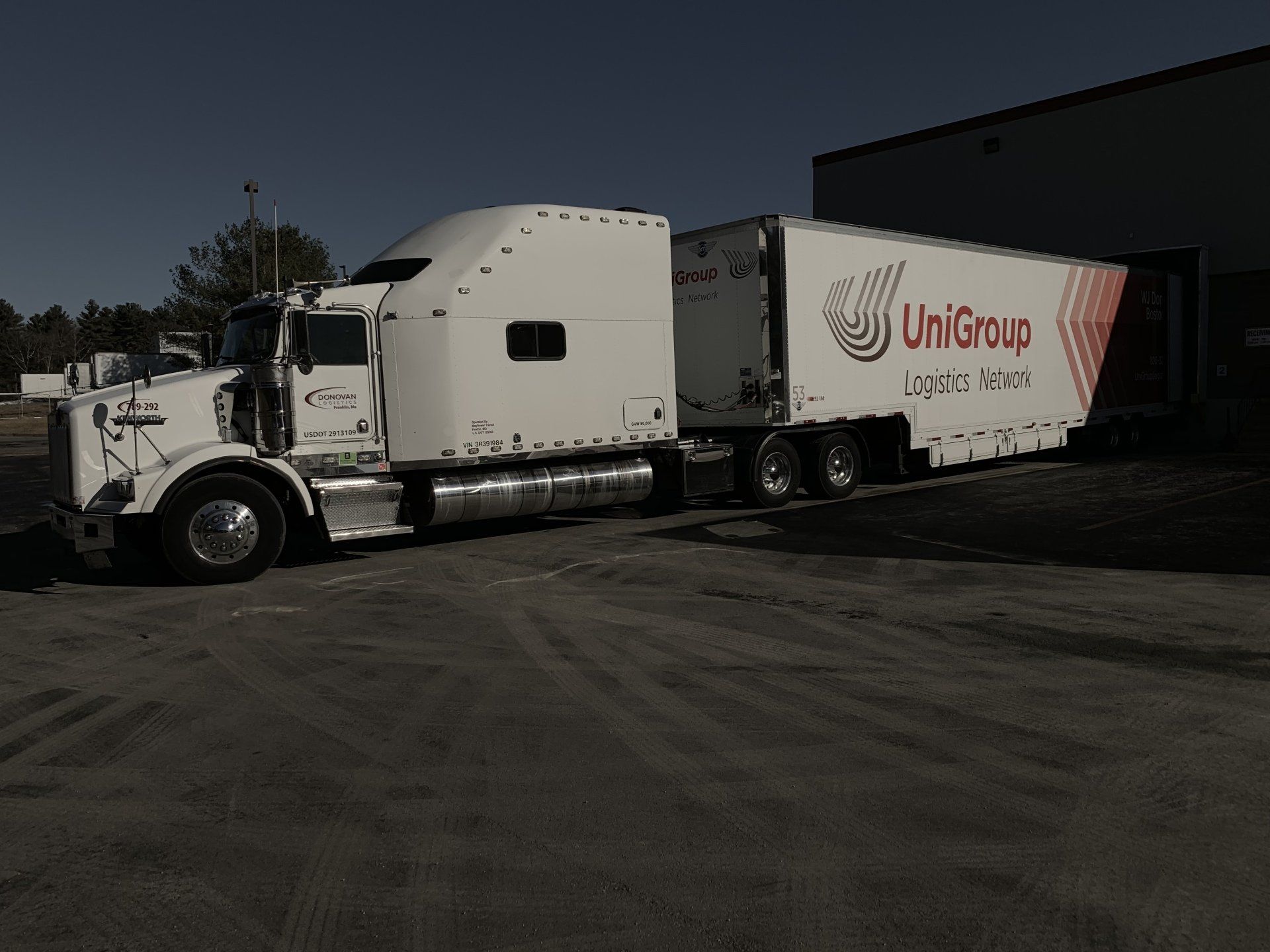 White semi-truck hauling a Unigroup trailer on a dark paved lot at dusk