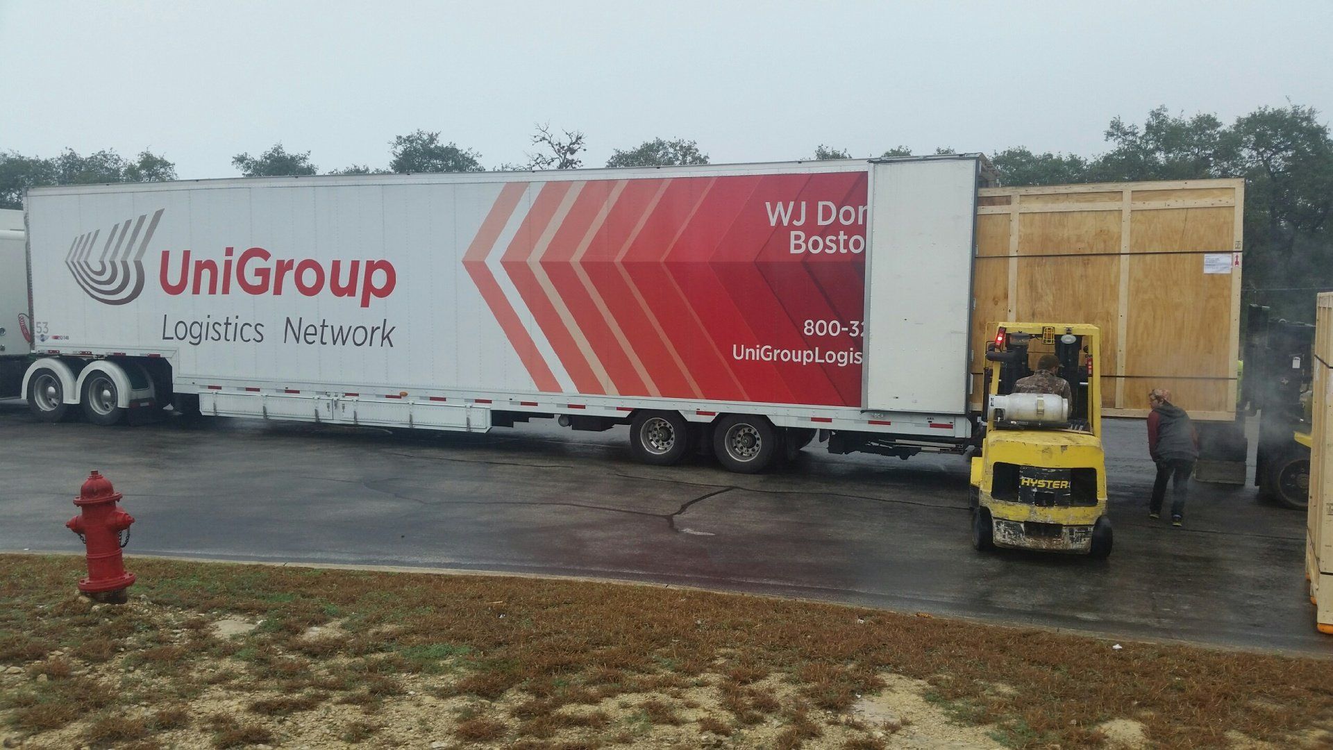 UniGroup moving truck parked by warehouse in rainy lot, with forklift and a red fire hydrant.
