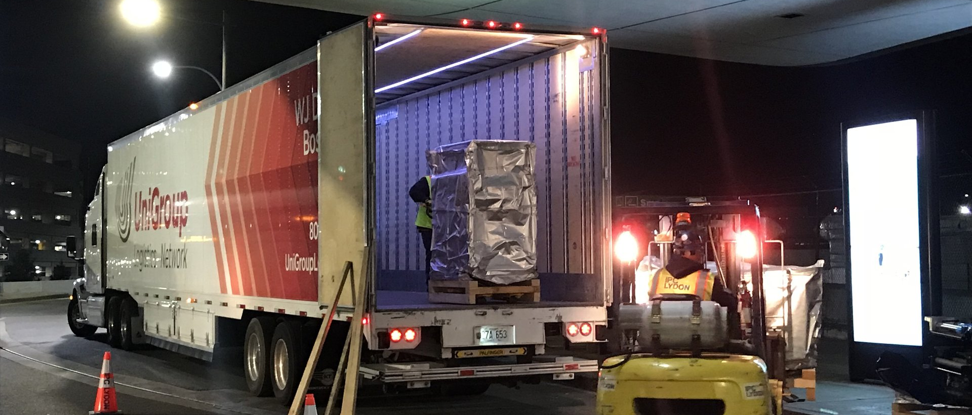 Nighttime dock loading of a truck container with workers and a forklift under bright lights