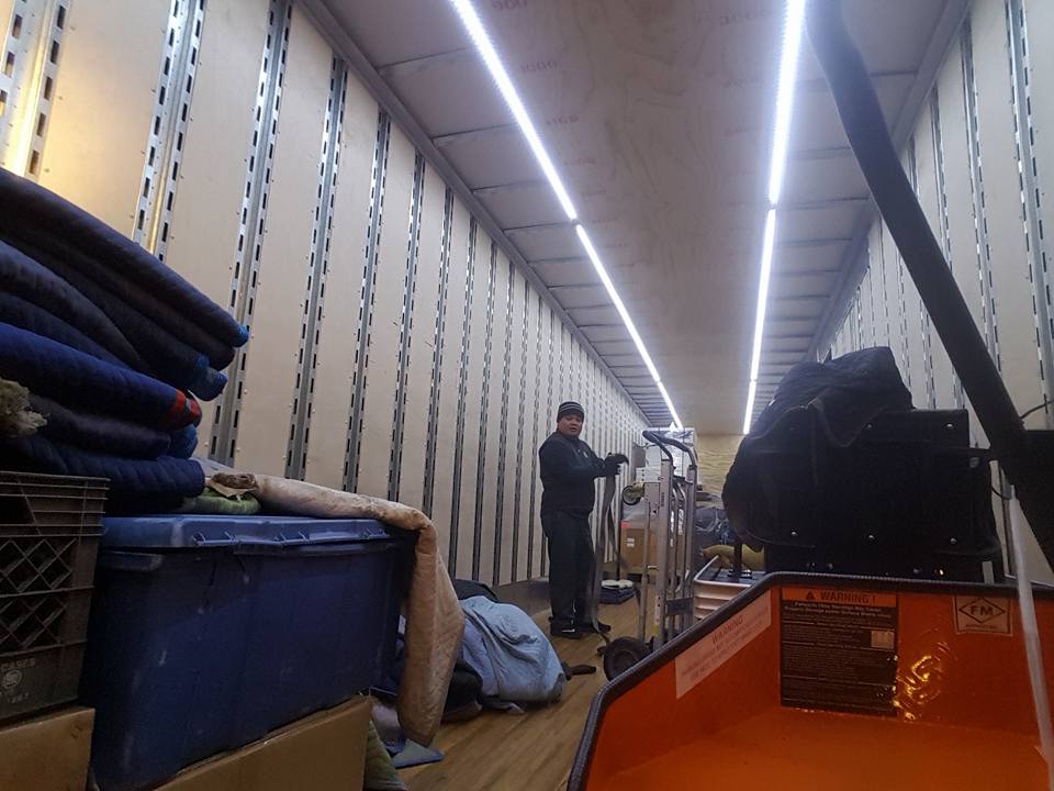 Person standing in a narrow warehouse aisle beside bins and stacked materials under bright overhead lights