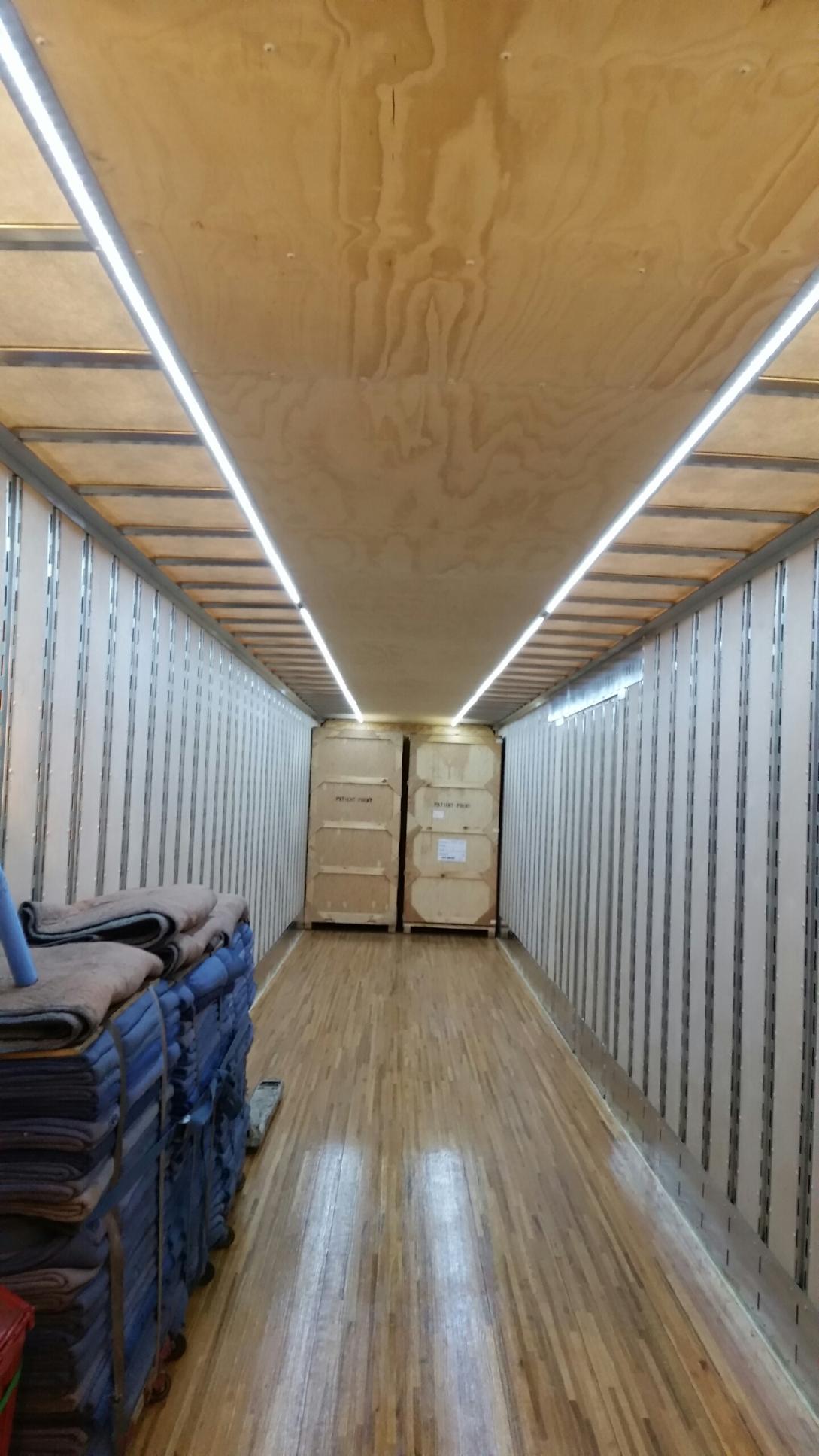 Inside a long cargo trailer, stacked folded items line the sides and boxes sit at the far end under wooden ceiling panels.