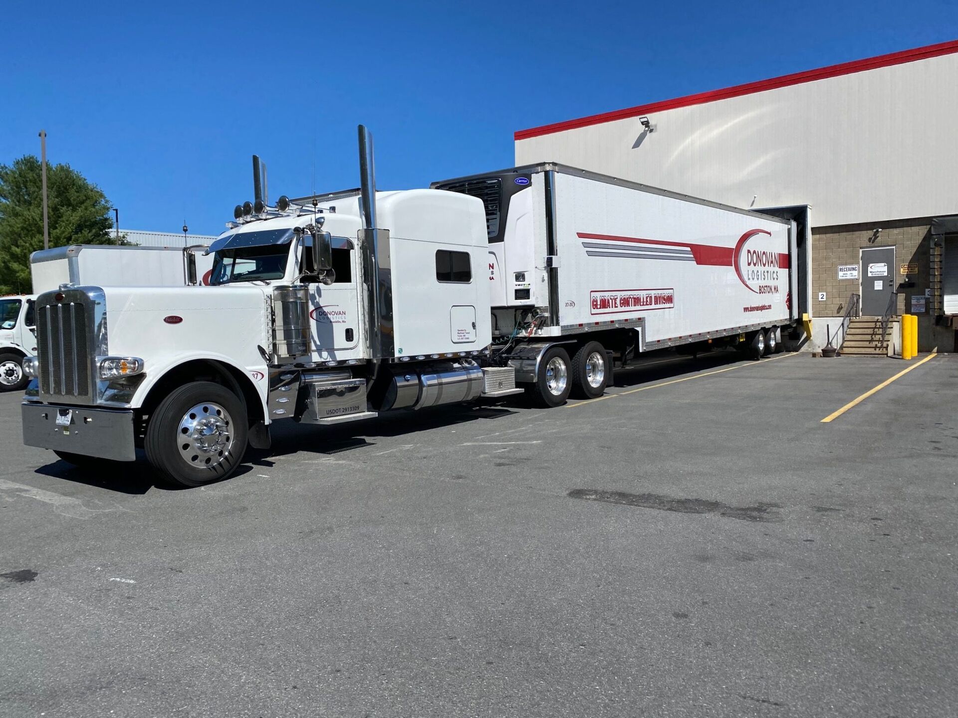 White semi-truck with a white trailer parked beside a warehouse loading dock on a sunny day