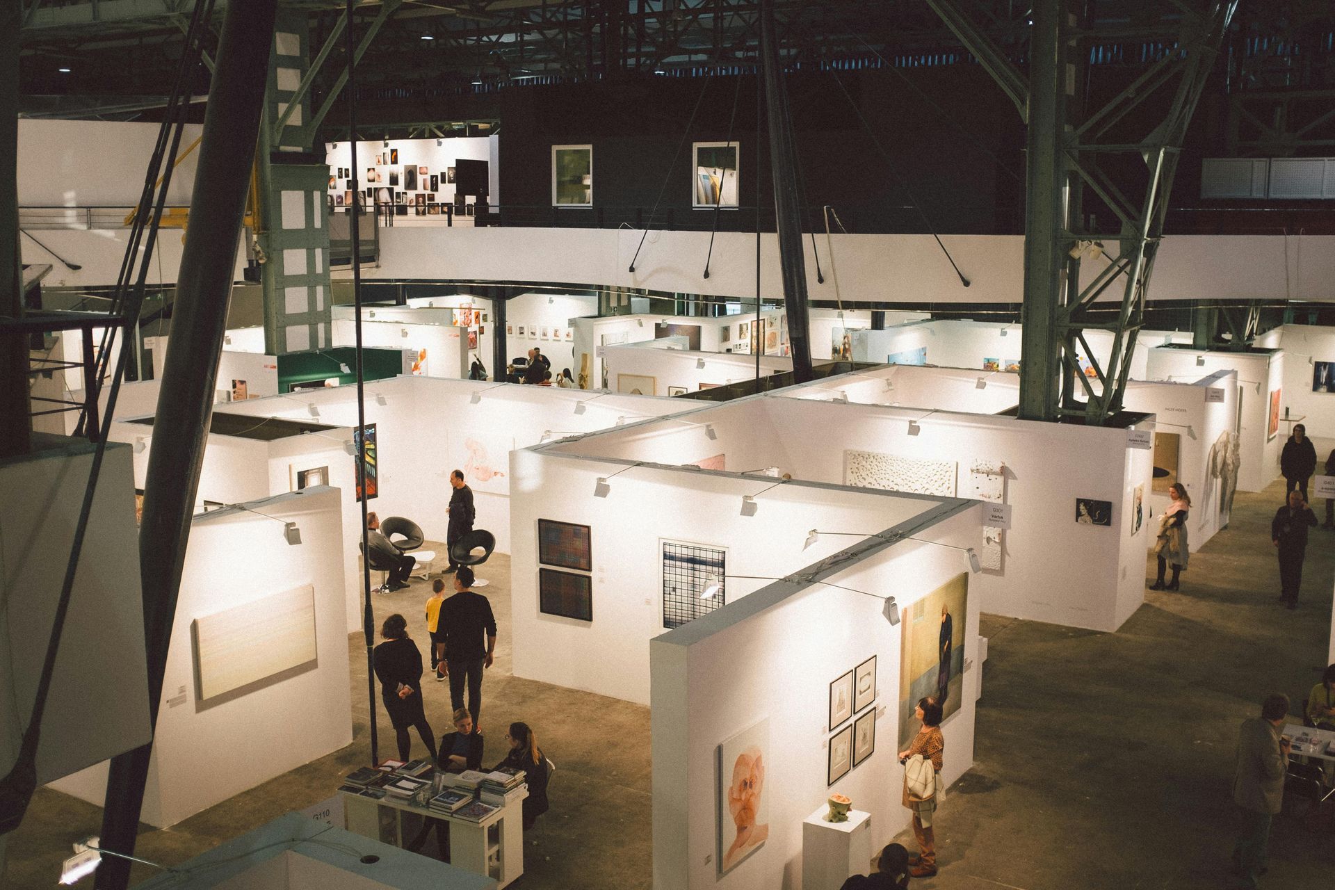 Indoor art gallery with white booths, framed artworks, and a few visitors under industrial lighting