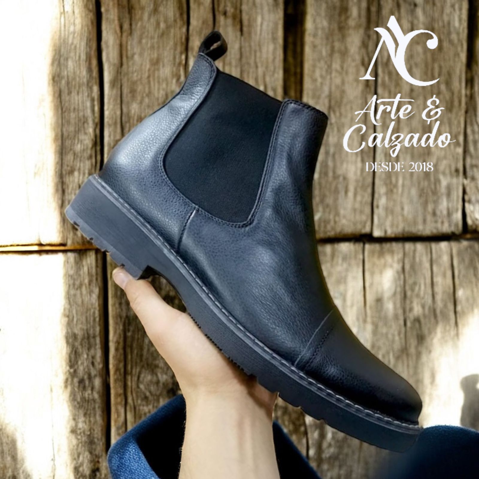 Una mano sostiene una bota Chelsea negra sobre un fondo de madera con un logotipo en la esquina superior derecha.