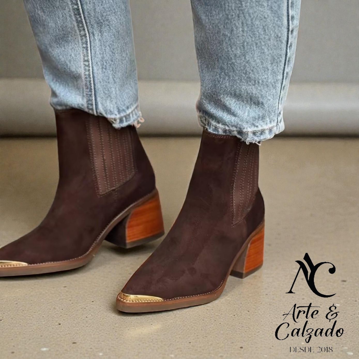 Botines Chelsea de ante marrón con tacón de bloque de madera y puntera dorada, combinados con vaqueros azul claro.