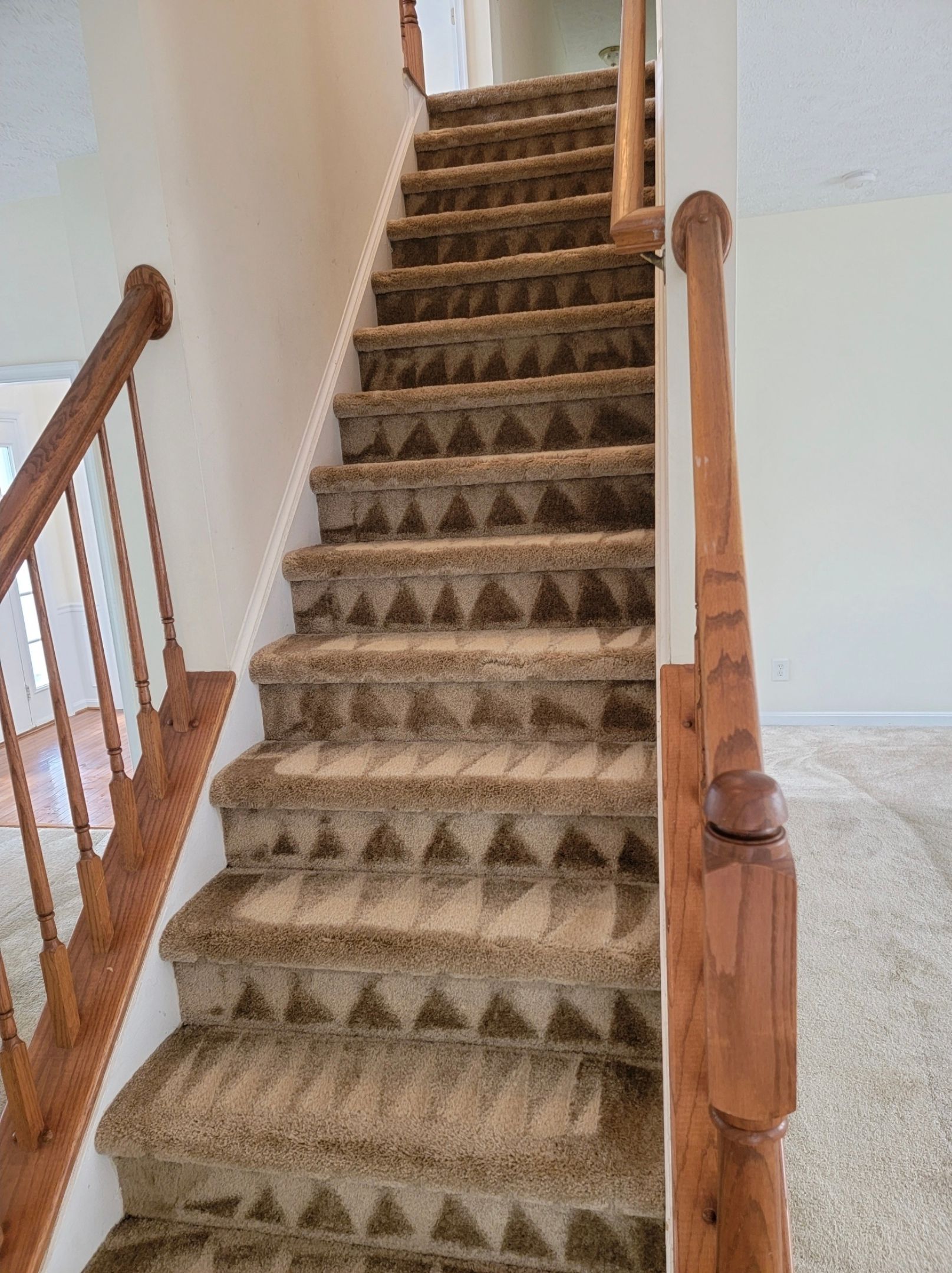 Staircase with brown carpet and wooden handrails leading upwards against a white wall.