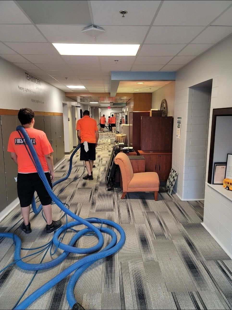Hallway flooded, people in orange shirts using blue hoses to remove water. Furniture sits along the side.