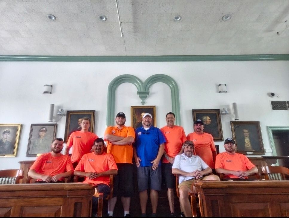 A group of people wearing orange and blue shirts stand in a room with wood paneling and portraits.