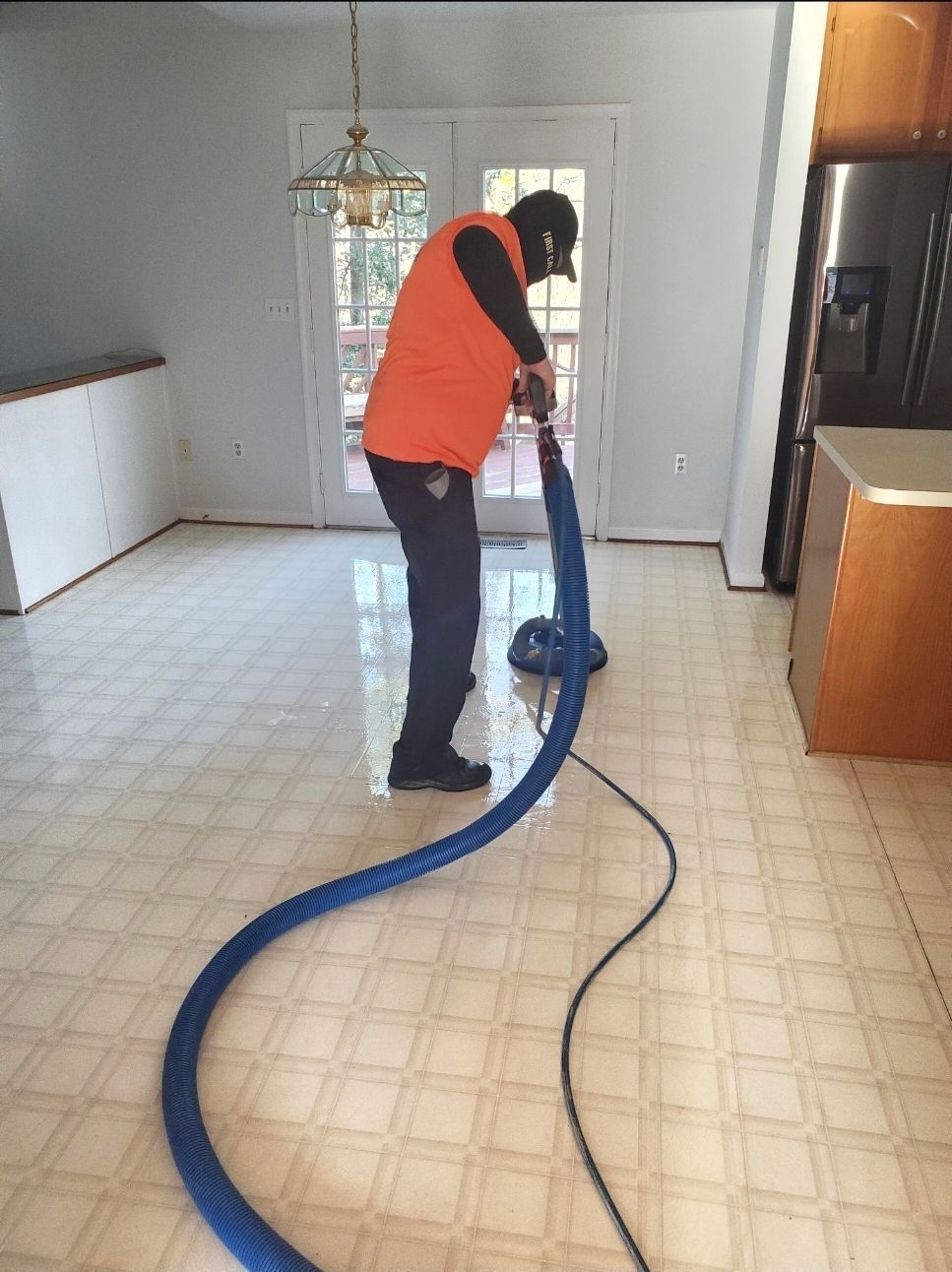 Person in orange vest cleans a vinyl floor with a machine in a kitchen.
