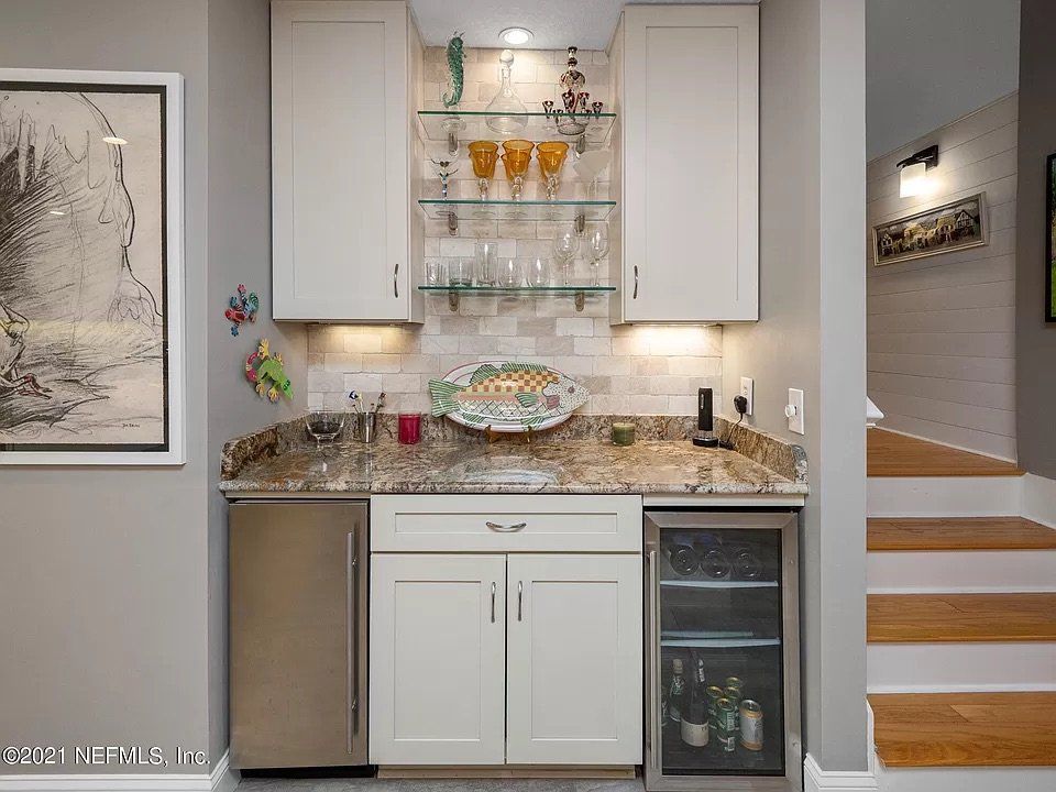 A home bar with white cabinets, granite countertop, mini-fridge, wine cooler, and glass shelves with glassware.