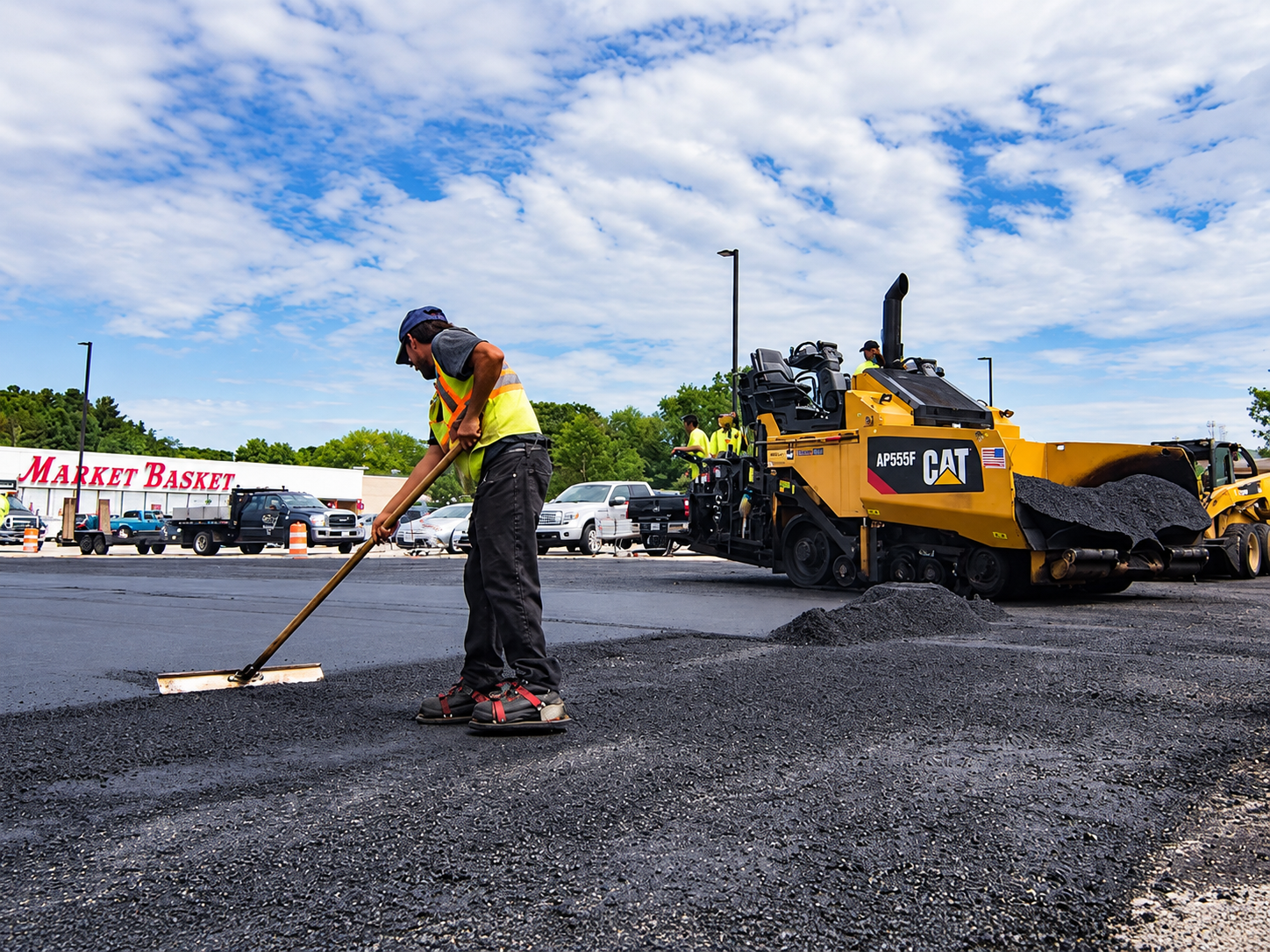 Worker rakes new blacktop beside the paver during Asphalt Paving Services in College Station, TX for a wide parking lot.