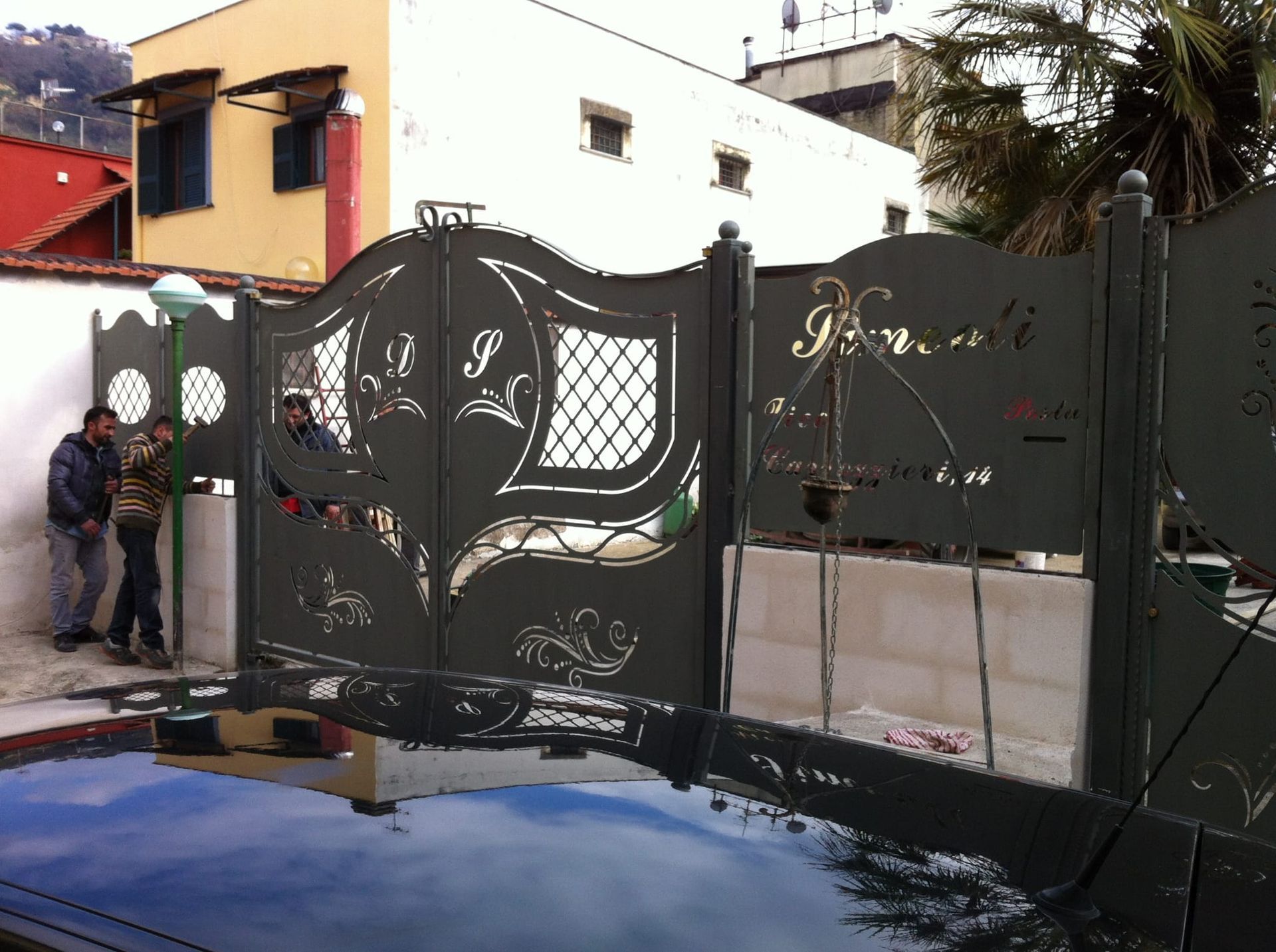 Cancello in metallo nero riccamente decorato davanti a una casa, con una persona in piedi a sinistra e il cofano di un'auto in primo piano.