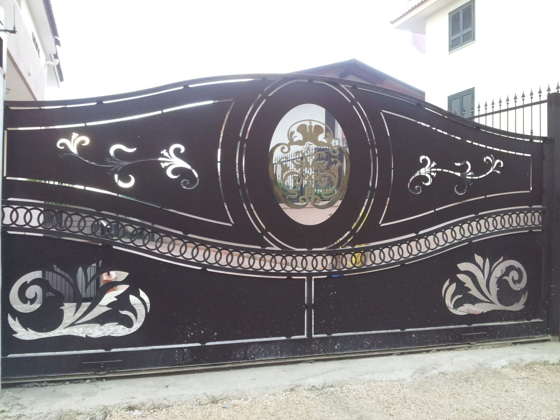 Cancello carraio in metallo nero decorato con motivi floreali bianchi e un emblema centrale ovale simile a uno specchio.