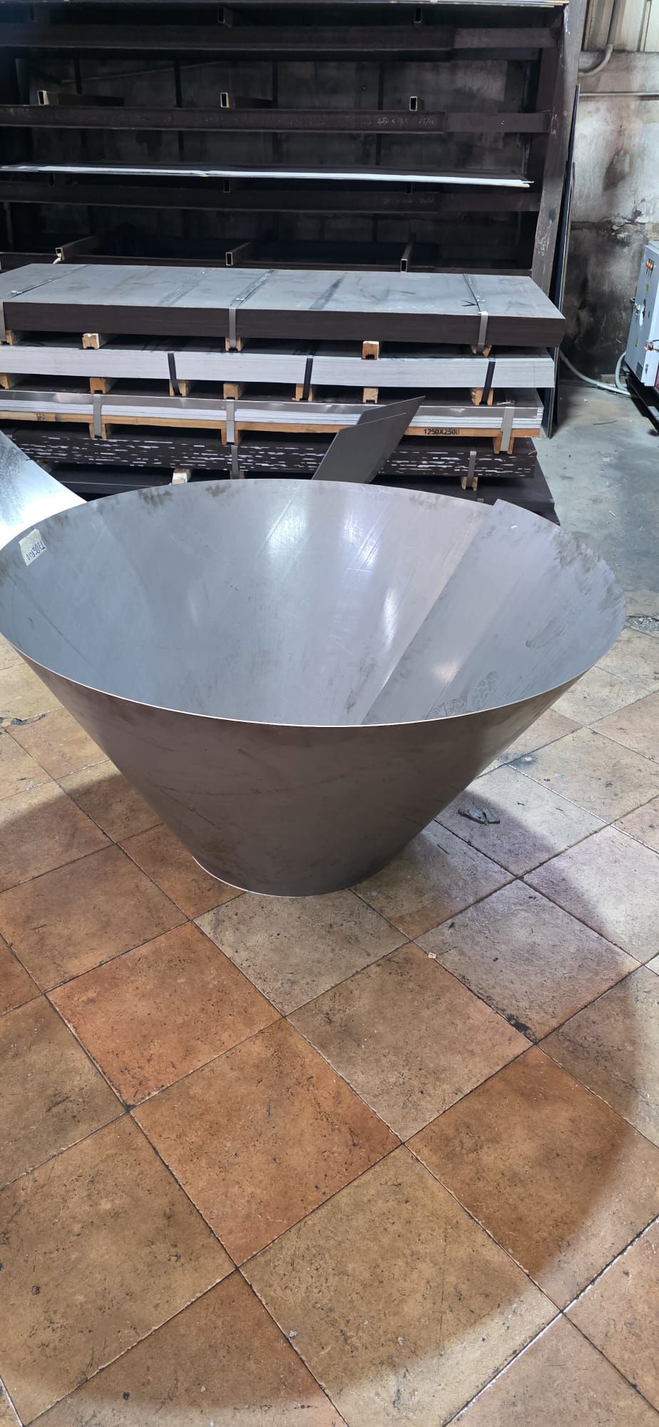 Grande braciere a forma di cono in metallo grigio su un pavimento piastrellato, con panchine e tavoli sullo sfondo.