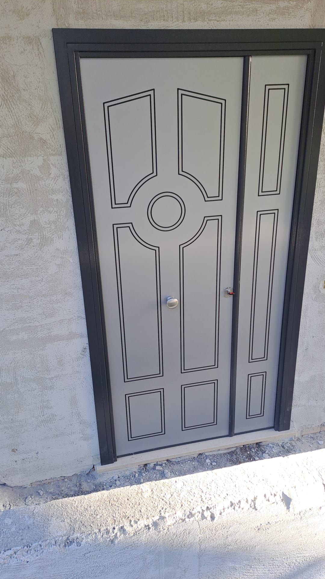 Porta d'ingresso in metallo grigio con pannelli decorativi e dettaglio centrale rotondo in un'apertura a muro con finitura materica.