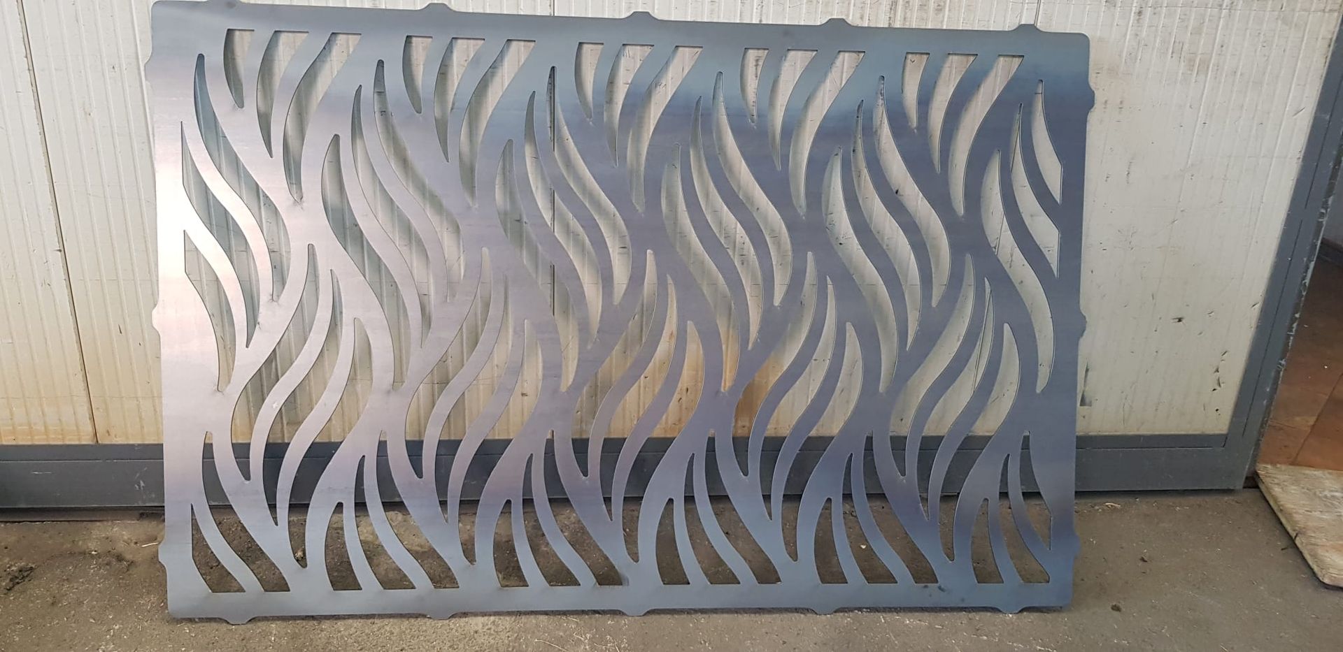 Pannello decorativo in metallo grigio con motivo a intaglio ondulato, appoggiato a una parete esterna.