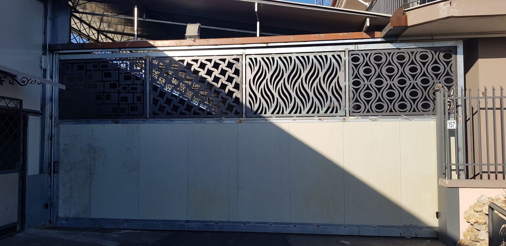 Cancello in metallo bianco con pannelli decorativi a griglia davanti a un edificio, parzialmente ombreggiato dalla luce del sole.