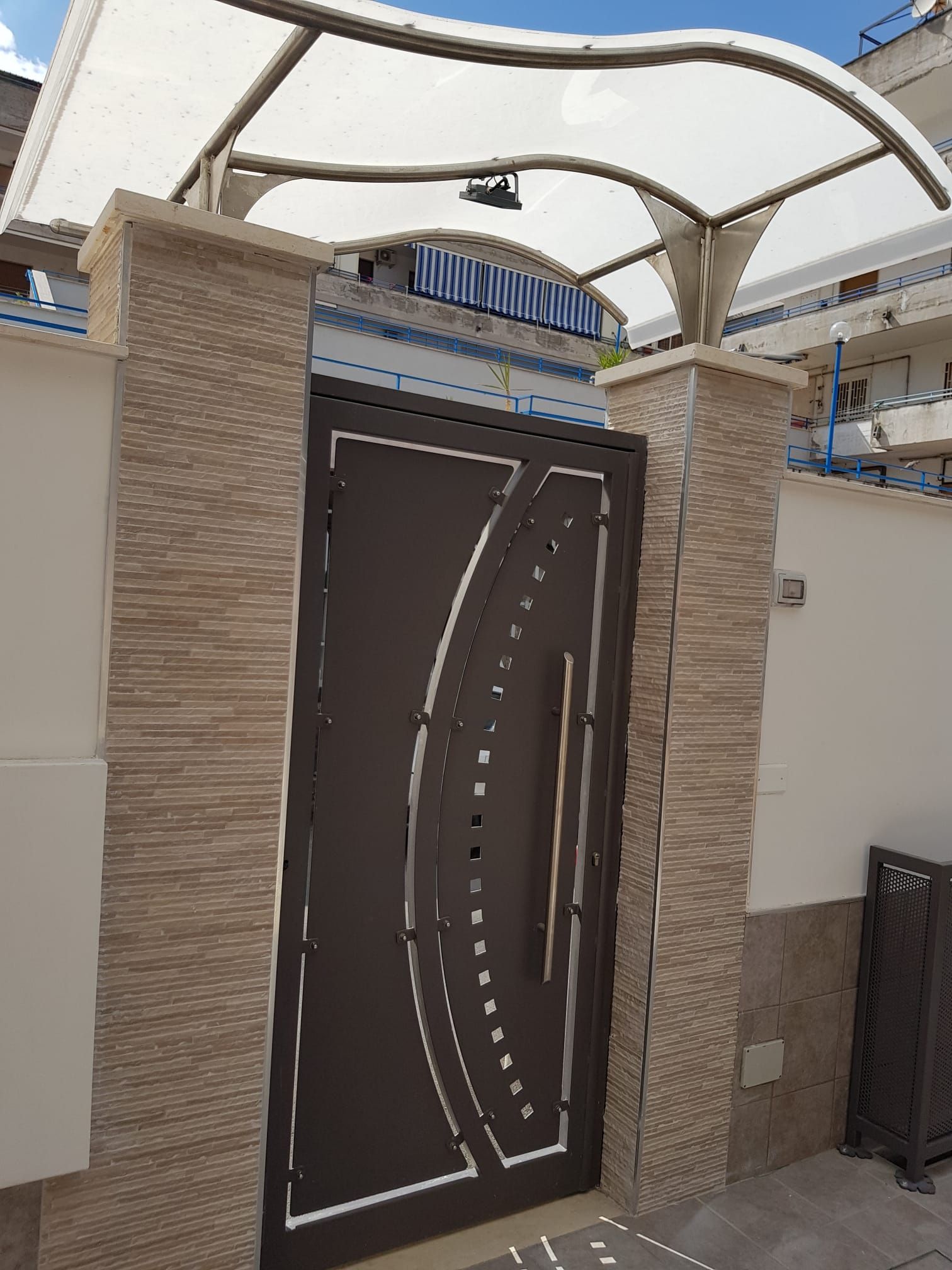 Cancello d'ingresso moderno con porta in legno scuro, pilastri in pietra beige e tettoia curva bianca in un cortile.
