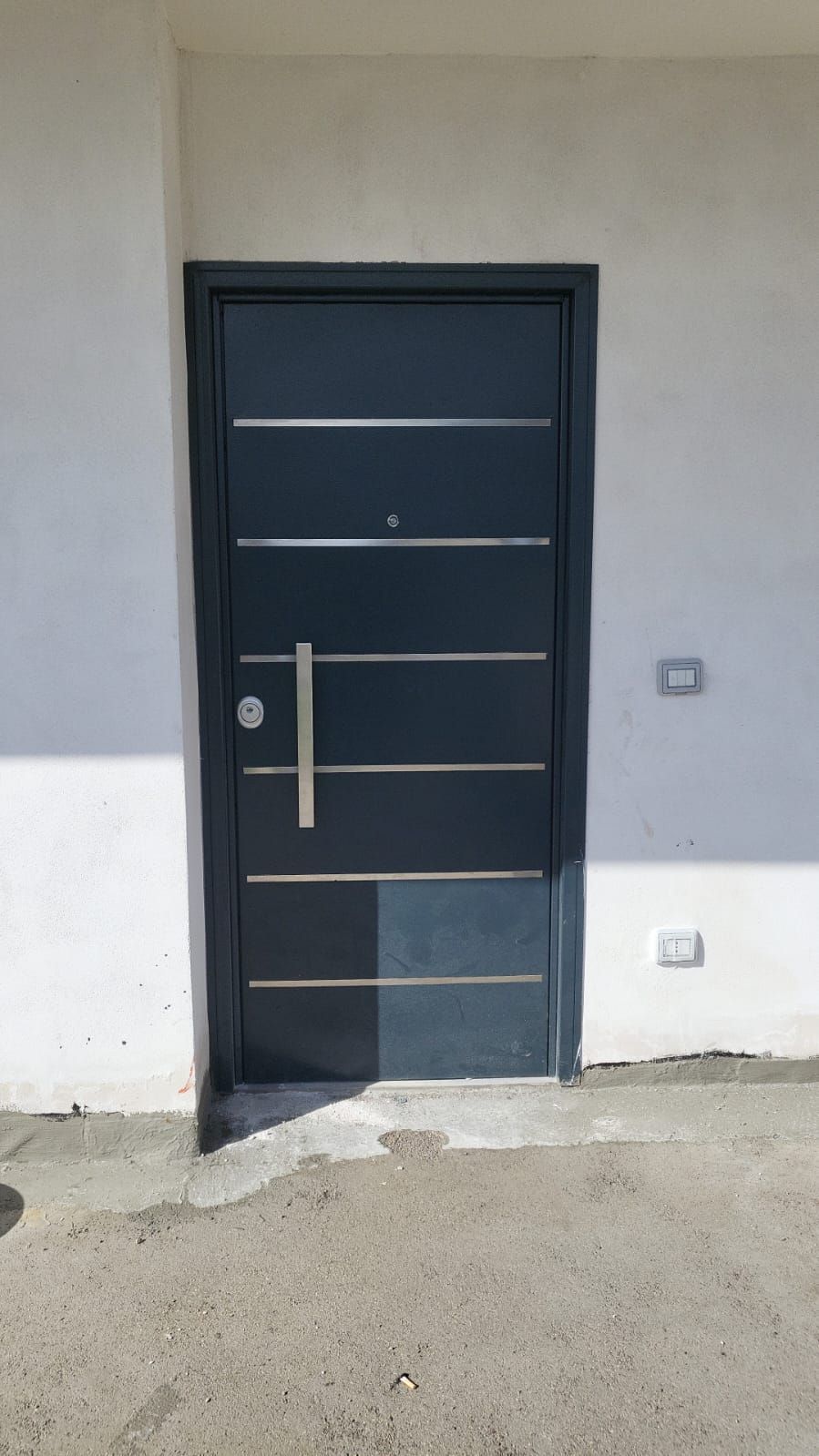 Porta nera con barre metalliche orizzontali incastonata in una parete grigia, vista dall'esterno su un pavimento di cemento grezzo.