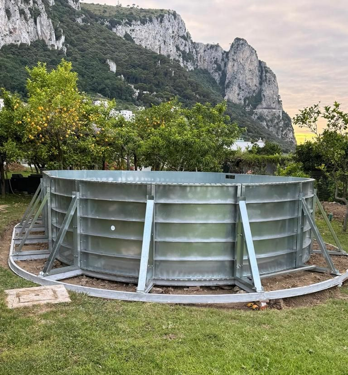 Una struttura circolare in acciaio zincato per una piscina fuori terra si erge in un prato ai piedi di una scogliera rocciosa.