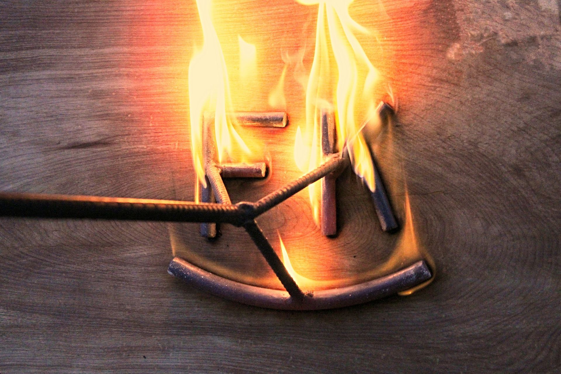 Flaming metal object on a wooden surface, with a dark rod touching it and bright orange flames rising.