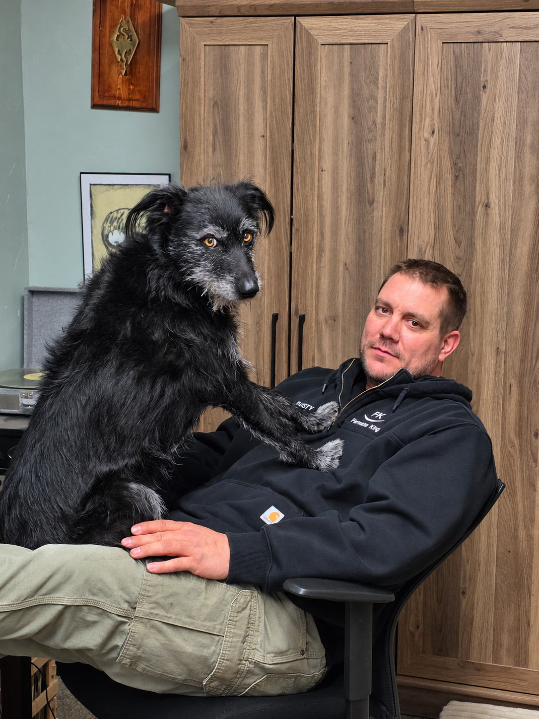 Man holding a large black dog on his lap in front of wooden cabinets