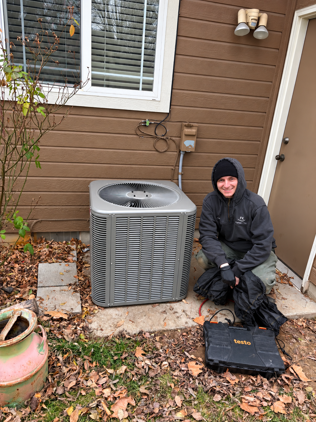 Person kneeling beside a gray outdoor air conditioner unit against a brown house wall