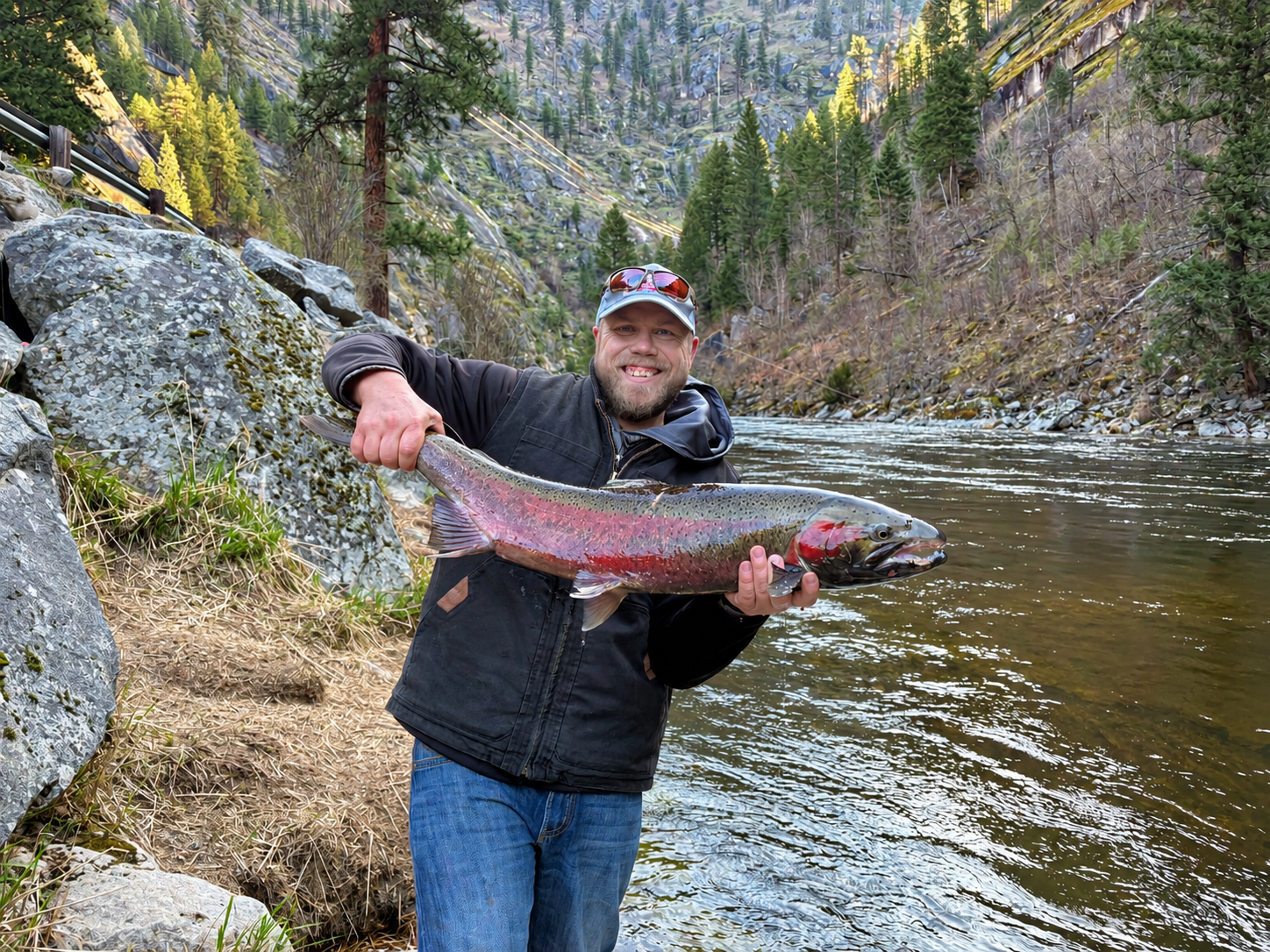 Man holding a rainbow trout beside a river in a forested canyon
