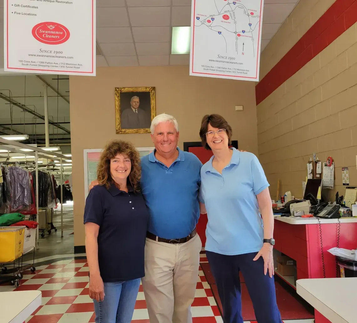 Three people stand smiling in a dry cleaners with red-and-white tiled floors, between two hanging signs.