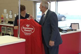 Santa Claus in red clothing stands in a shop, holding a branded garment bag from Swannanoa Cleaners and giving a thumbs-up.