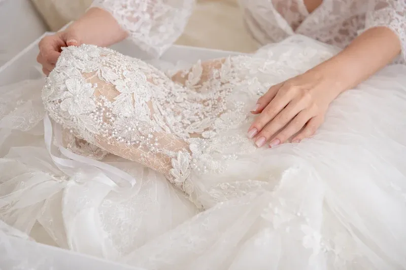 Hands gently touch an intricate white lace wedding dress as it is laid out inside a box.