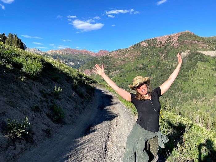 Erika stands with joyfully widespread arms, presenting the mountains behind her.