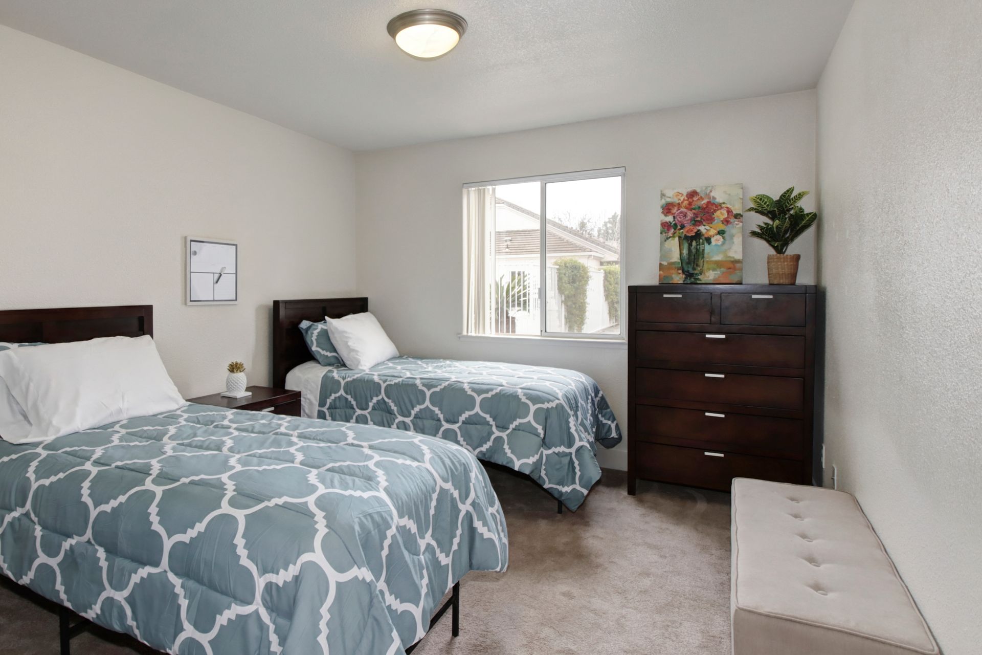 Bedroom with two twin beds, dresser, and a window. Blue patterned bedding, neutral walls, and a small bench.