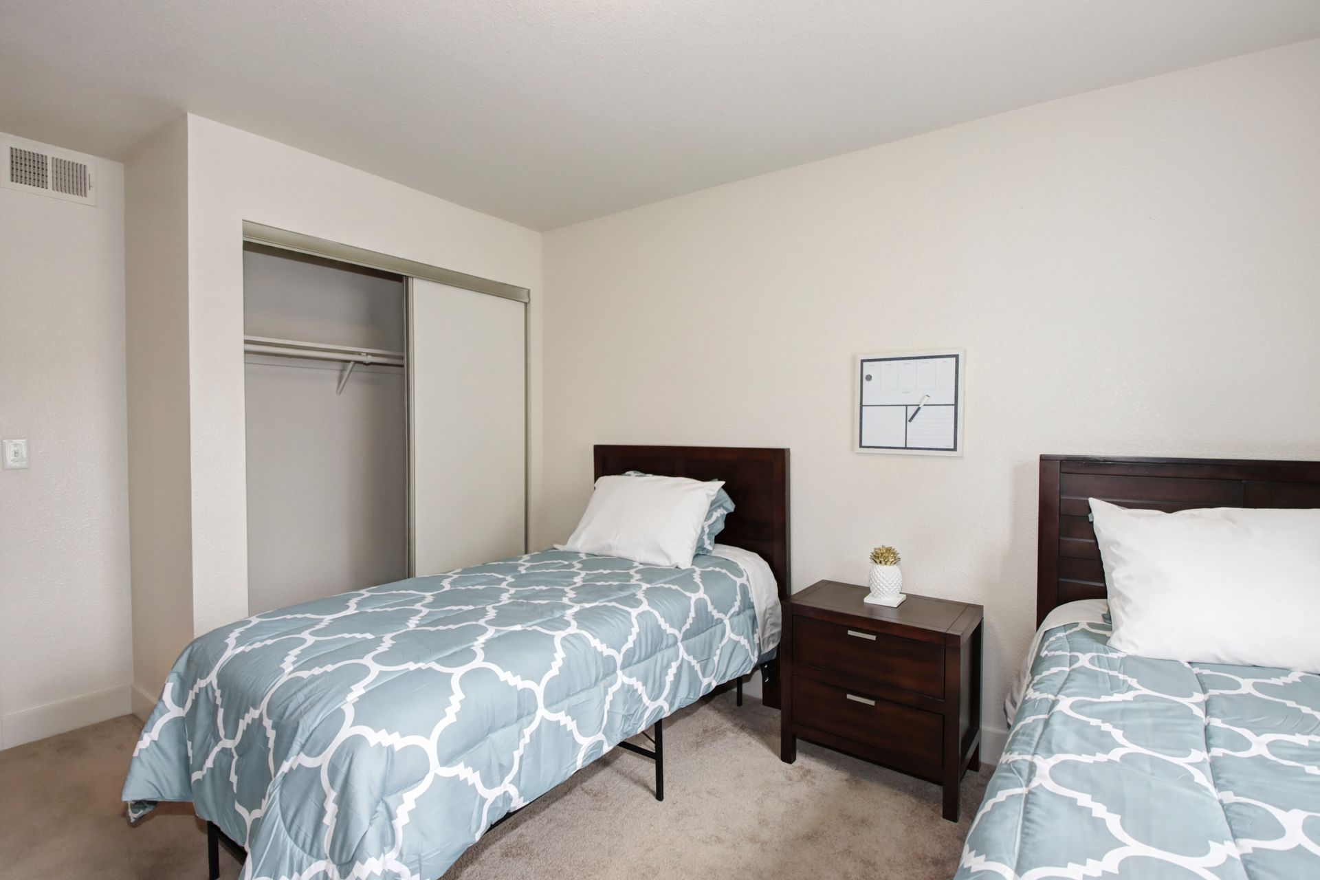 Two twin beds with blue and white bedding in a beige-walled room. A nightstand sits between the beds.