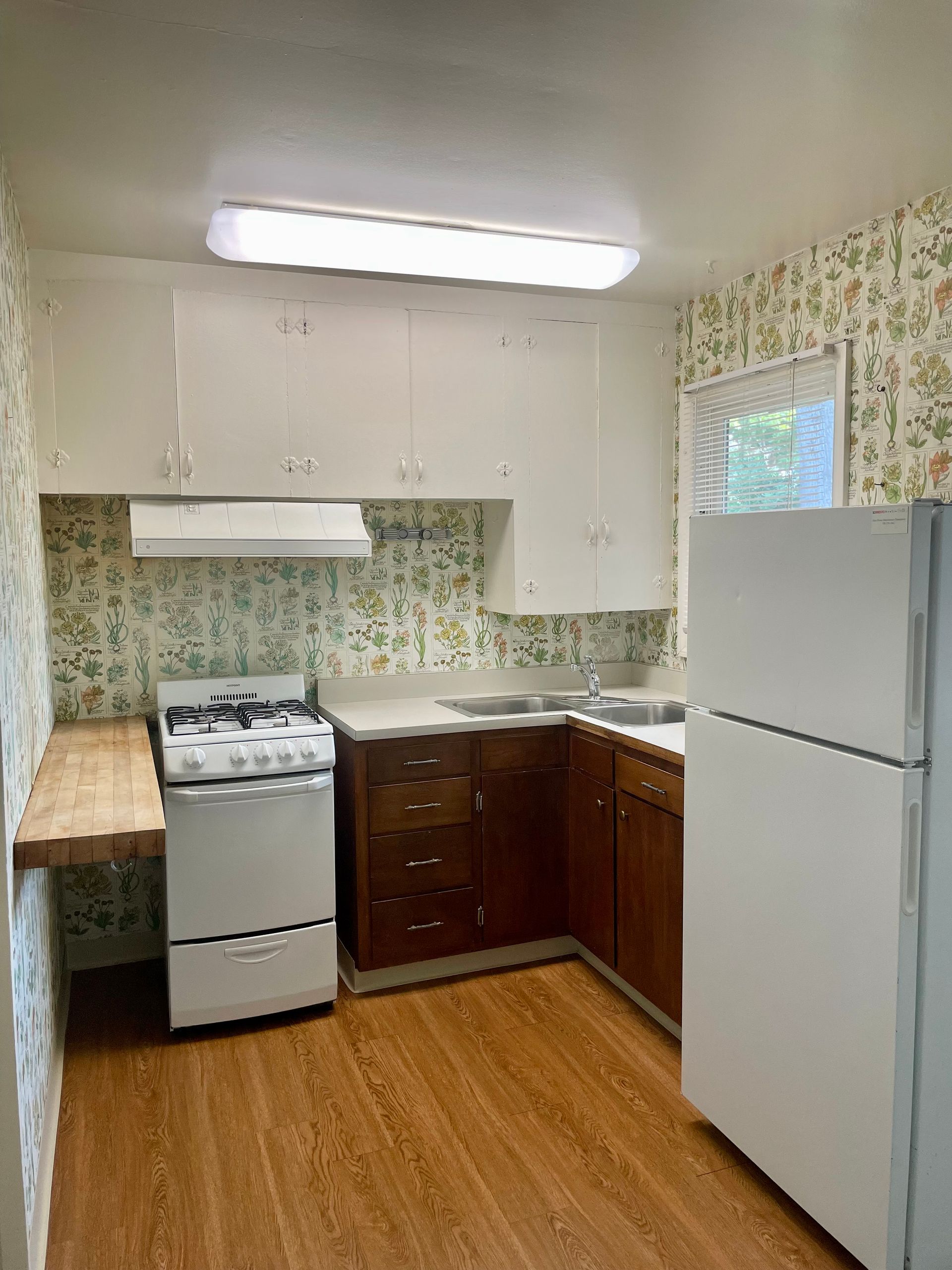 Small kitchen with wood cabinets, floral wallpaper, white appliances, and a wood-look floor.