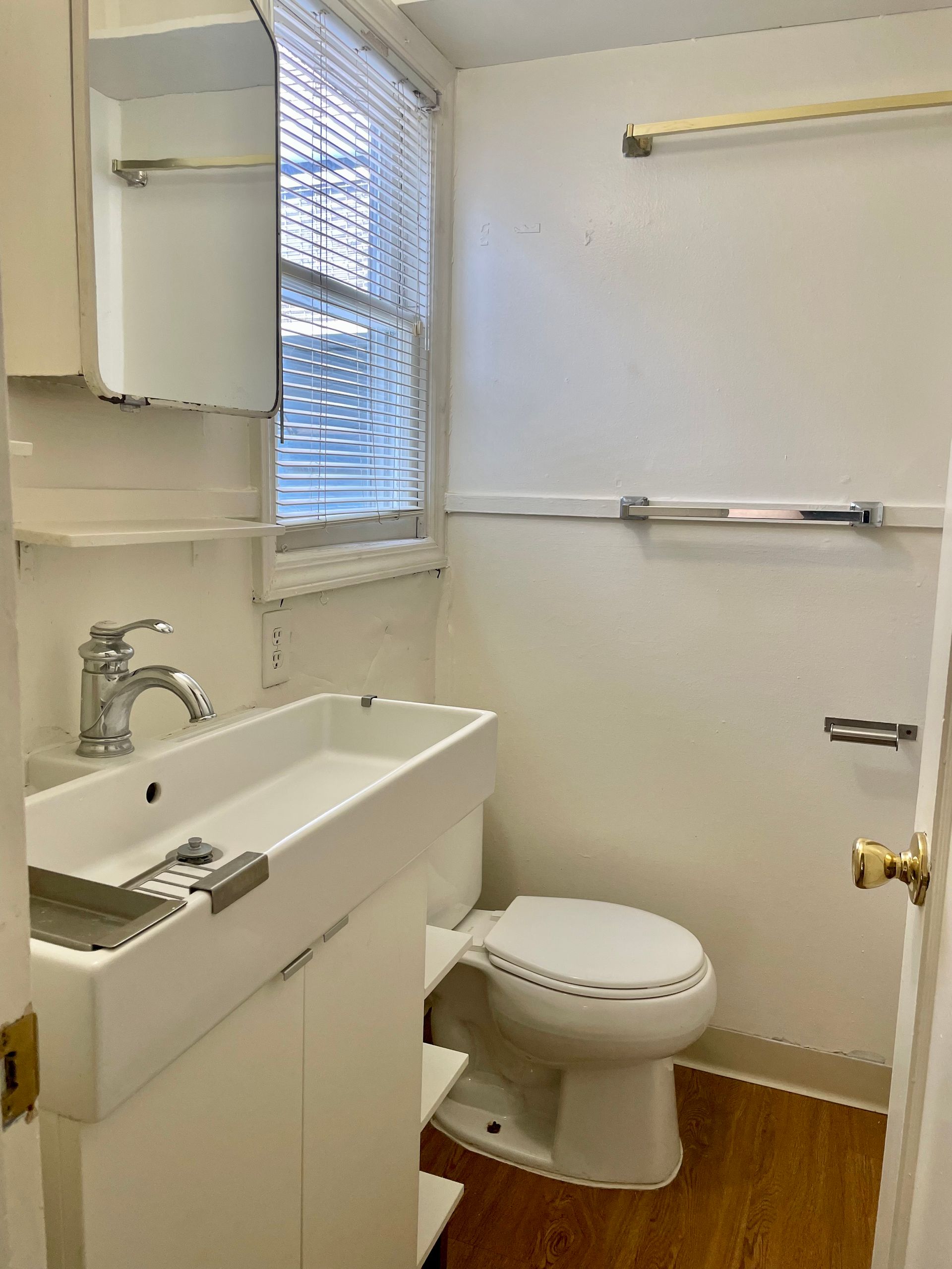 Small white bathroom with toilet, sink, window with blinds, and a cabinet.