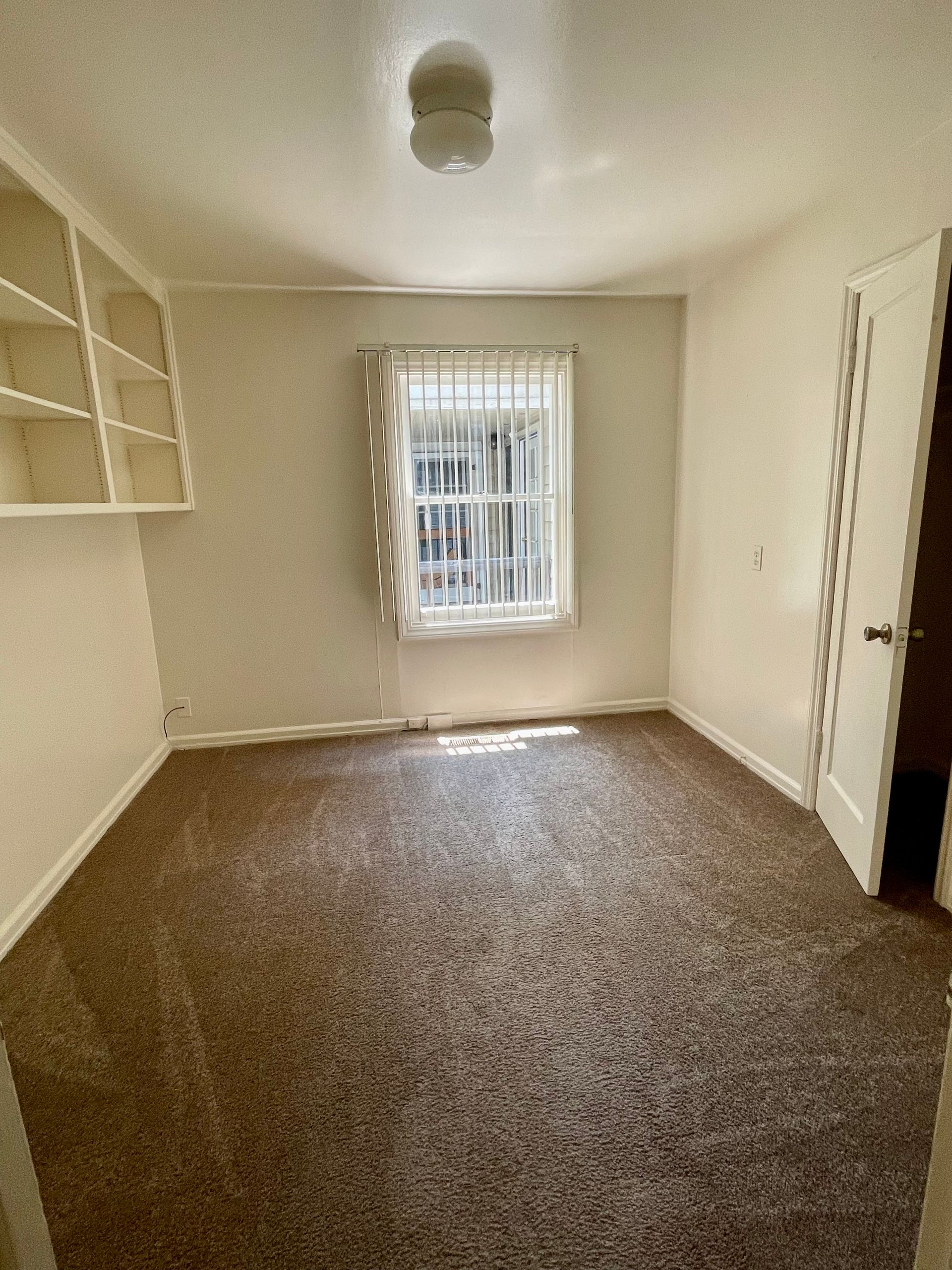 Empty room with brown carpet, a window with blinds, and built-in shelving.