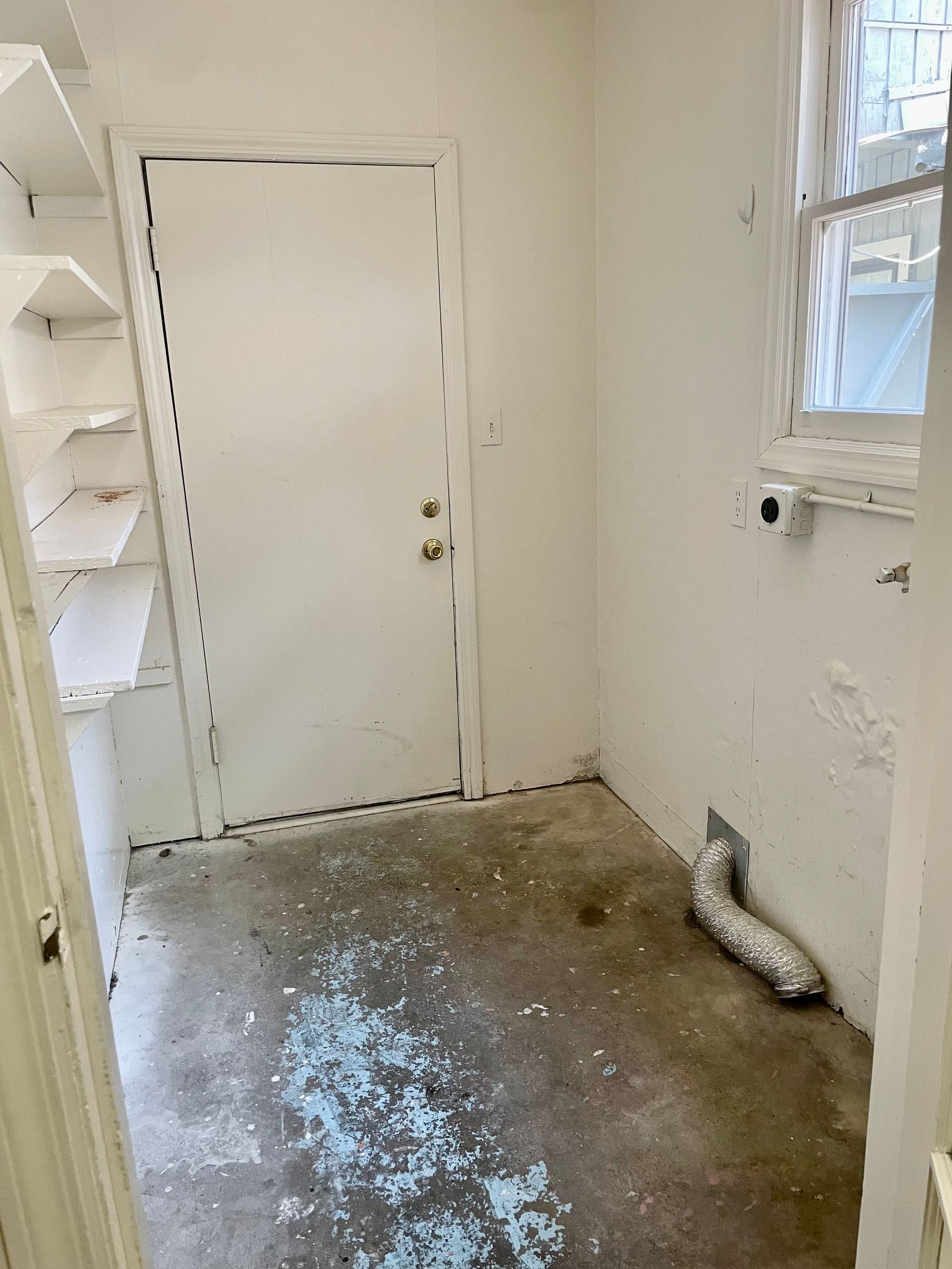 A small utility room with a door, shelves, and a window. The floor is stained, and the walls are white.