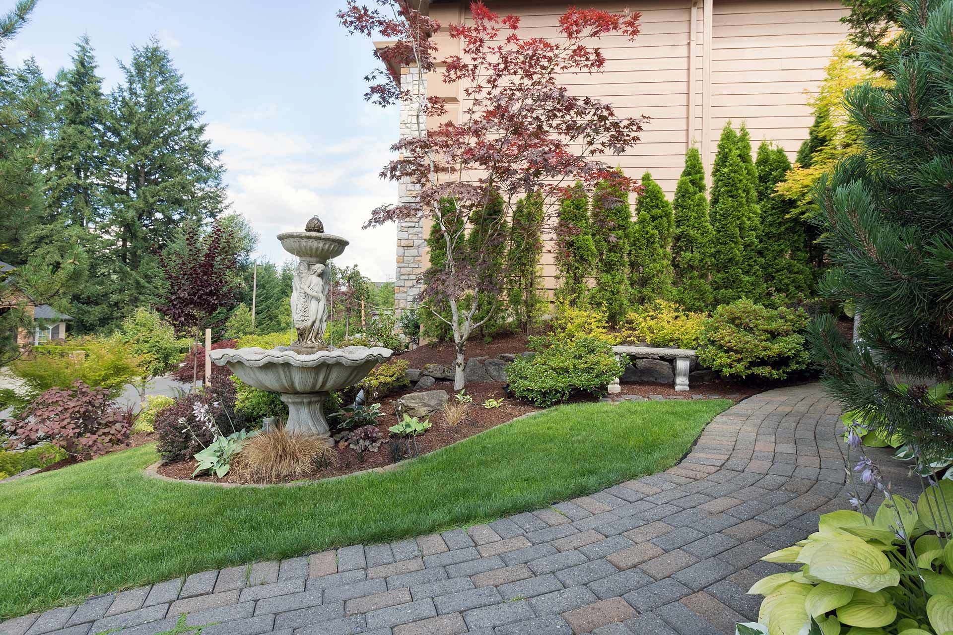 Landscaped garden with fountain, curved stone path, shrubs, and a house facade