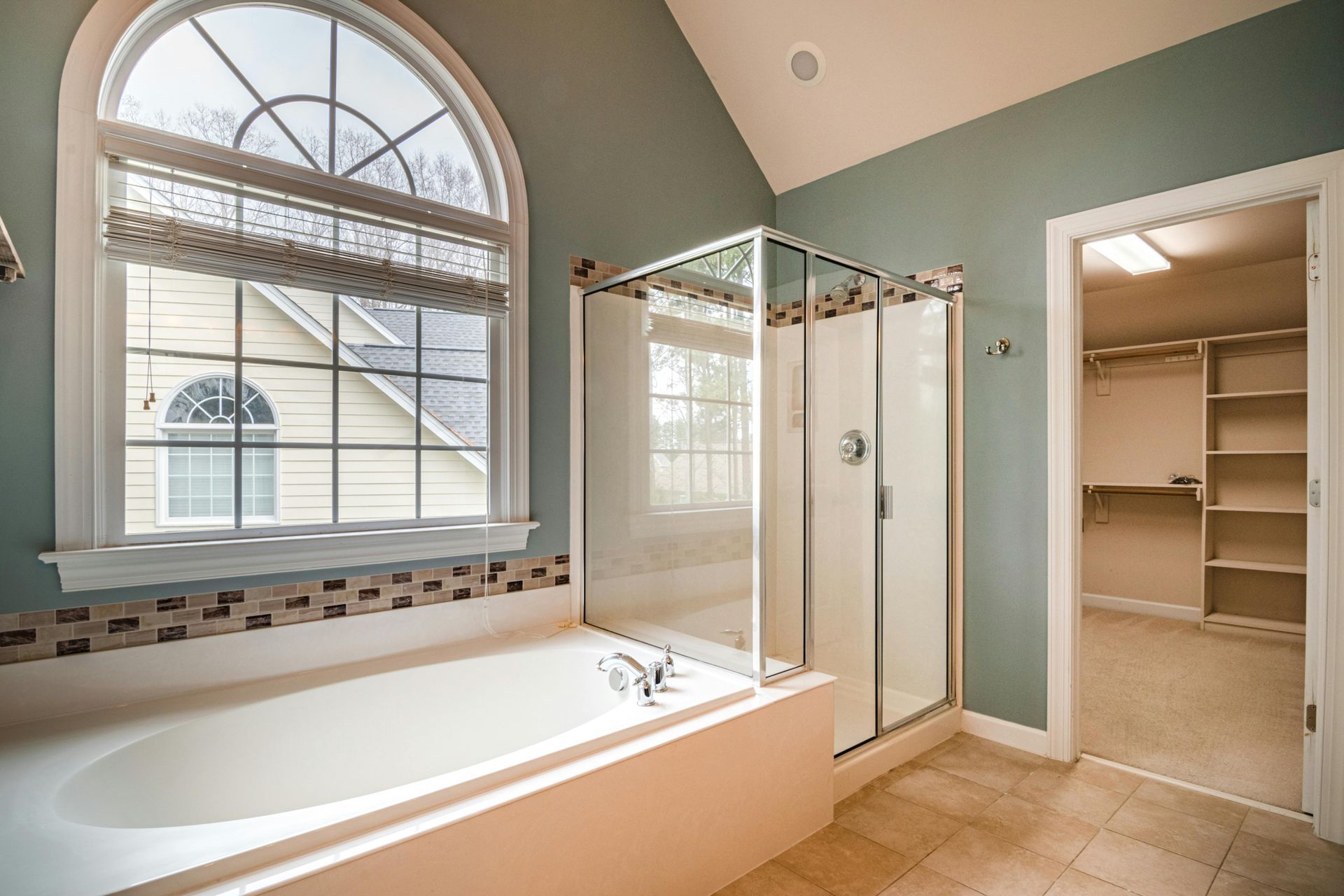 Bathroom with big white bathtub connected to a glass door walk in shower. It has green paint, a dark brown backsplash, light brown tile, white crown molding, and a huge walk in closet.