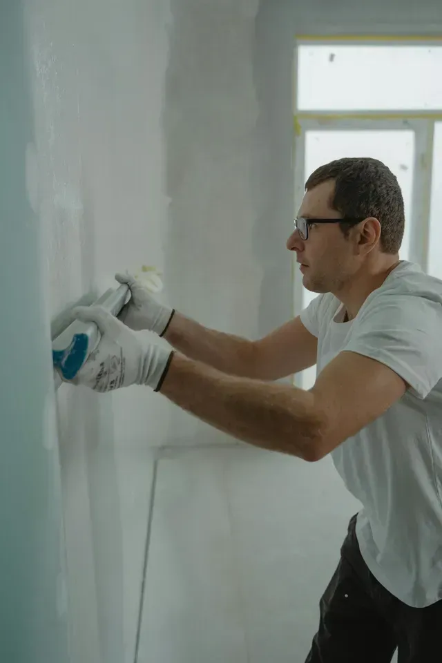 A man with a white T-shirt, blue jeans, and glovesis working on drywall in a room in a house.