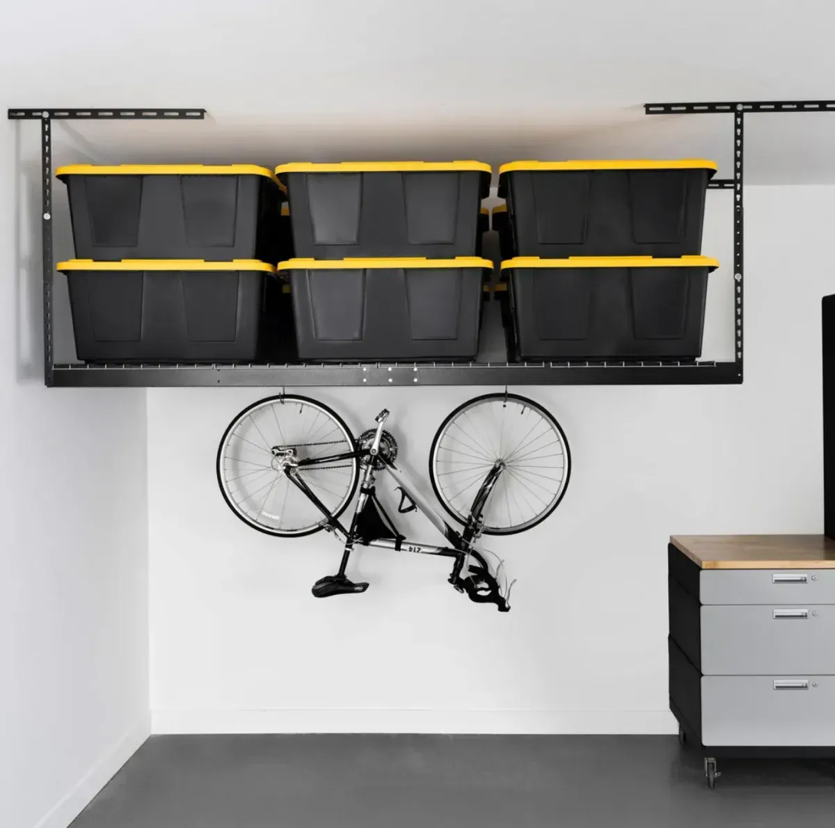 A large black ceiling storage rack hanging in a garage. One mountain bike is hanging from it upside down, and six black bins with yellow lids are stored on the rack.