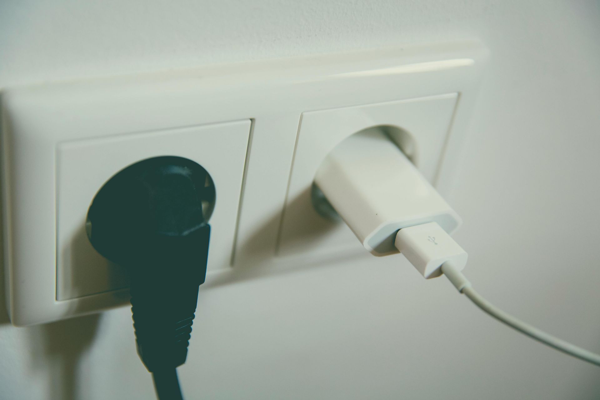 Standard duplex 15 amp outlet with one black cord and one white phone charger plugged in.