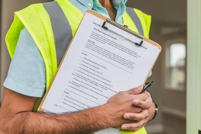 A maintenance technician wearing his yellow reflector vest is holding a clipboard with a checklist.