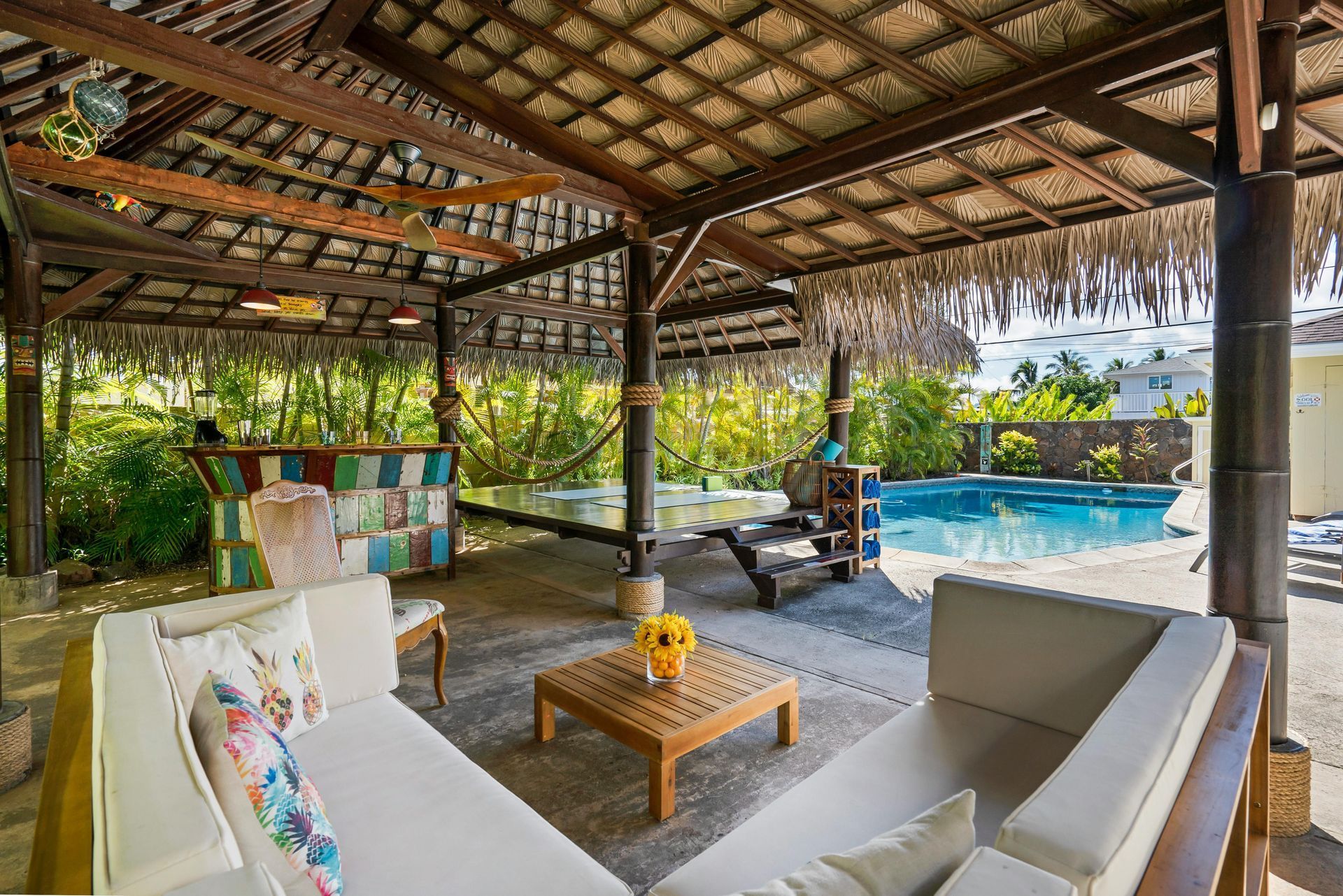 Beautifully done backyard with a wood beach style thatch  cabana with ceiling fans and pendant lighting, outdoor patio furniture with white cushions, brown stamped concrete, a small wading pool, and small turquoise and brown colored bar.