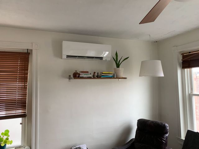 White AC unit on a wall above a shelf with books and a plant. Brown blinds and windows frame the scene.